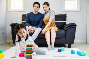 Happy asian family father and mother with little asian girl smiling playing with building tower from wooden blocks, taking tiles in turn from underneath until it falls, in moments good time at home photo