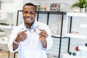 Eyesight And Vision Concept. Portrait of smiling african american standing at optics store and pointing at showcase and rack with many eyewear and specs frames. Guy offering spectacles photo