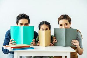 Cloesup portrait of adorable attractive beautiful asian family, Asian young mother and father with little daughter sit at desk and holding book with funny facial expression photo