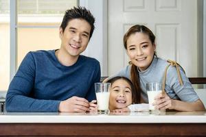 Happy family having fun in the kitchen. Asian Father, mother and little daughter spending time together and having breakfast drinking and hold glasses of milk at table photo