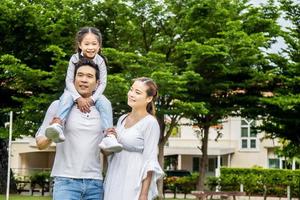 Portrait of asian happy family of three having fun together in park,  little daughter sitting on father back and mother smile walking and having fun moments good time in summer park at home photo