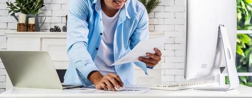 Young businessman smiling happy asian man working holding a piece of paperwork and reading while standing in front of laptop at home, work from home concept photo