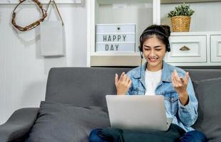 Asian young business woman is sitting on the sofa in her own house is wearing headphones video call conference with laptop computer raise hand saying hi team at home office, work from home photo