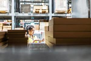 Asian female worker in safety vest sitting and working with computer laptop in storage warehouse. people, warehouse and industry concept photo