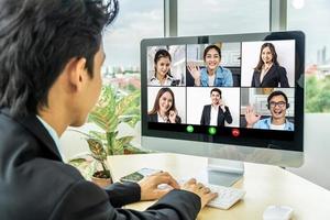 Business man sit at desk looking at computer screen where collage of diverse people webcam view. Asian man lead video call distant chat, group of different mates using video conference photo