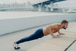 Horizontal shot of muscular athlete man does push up exercise has strong body stands in plank pose outdoor wears sport trousers and sneakers concentrated into distance. Strength and motivation concept photo