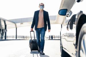 Travelling guy pulls suitcase, resturns from airport, approaches car, wears medical mask to prevent coronavirus, takes taxi from airport. Dangerous travel, influenza, quarantine and epidemic concept photo