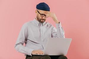 Male entrepreneur tries to make solution, keeps hand on head, tries to prepare financial report, focused in laptop computer, poses against pink background. Unshaven young man browses website photo