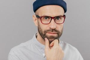 Close up shot of handsome bearded young man holds chin, looks seriously directly at camera, thinks about something, wears spectacles, white clothes, isolated over grey background. Indoor shot photo