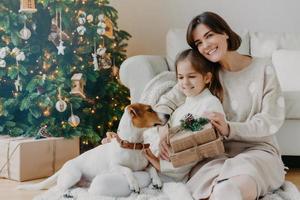 felices vacaciones de invierno. mujer morena positiva abraza la pose de la niña con regalos en el piso de la habitación, el perro jack russell terrier cerca, diviértete cerca del árbol de navidad. mamá e hija desempacan cajas foto