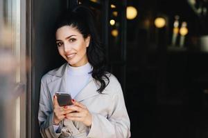 mujer morena sonriente con cola de caballo con manicura roja vestida formalmente sosteniendo un teléfono inteligente haciendo compras en línea mientras espera el tren. mujer de negocios con teléfono celular yendo a trabajar foto