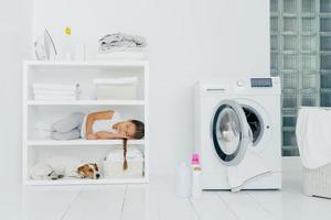 Indoor shot of little girl has sleep on console with favourite dog, has rest in laundry room with washing machine filled of linen, bottles with detergent. Children, tiredness and house work.
