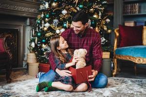 Small female child holds teddy bear, recieves unexpected gift from affectionate father, being thankful to him. Young dad gives present to daughter, congratulates her with Christmas or New Year photo