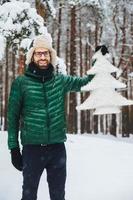 el retrato de un alegre y apuesto hombre barbudo lleva un sombrero caliente, un anorak verde y guantes sostiene un pequeño abeto artificial blanco, se encuentra en un bosque invernal cubierto de nieve. hermosos paisajes foto