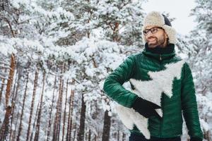 Cheerful pleasant looking man with beard looks thoughtfully aside, holds fir tree, wears spectacles, hat and jacket, poses against winter trees covered with snow. People and lifestyle concept photo