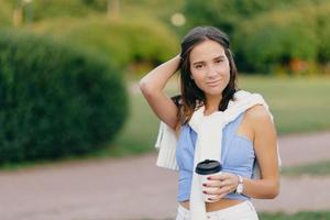 Pleased beautiful European woman with fit figure, keeps hand on head, drinks takeaway coffee, stands against green blurred background with copy space for your advertisement. Coffee time concept photo