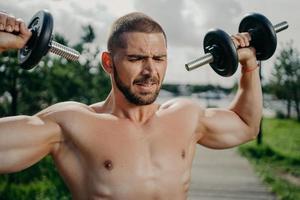 Sporty determined muscular European man exercsises with dumbbells, makes weightlifting outdoor, has naked torso, trains muscles, wants to have athletic body. Strength and motivation concept. photo
