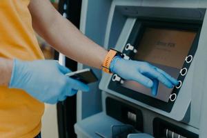 Cropped shot of man wears protective rubber gloves, uses ATM machie to withdraw money, prevents from corona virus or covid-19, holds mobile phone, presses button. Virus pandemic. Health care concept photo