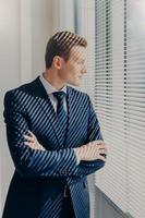 Waist up portrait of handsome male financier looks attentively in office window, has thoughtful expression, rests after conference, has work break, keeps hands crossed, wears elegant clothes photo