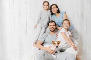 Horizontal shot of friendly family pose together against white background two little sisters, father, mother and their pet. Happy parents and their female children. Family of four. Parenthood photo