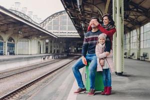 Glad female covers eyes of her husband, makes surprise. Friendly family of mother, father and daughter stand together at railway station, have fun while wait for transport. People and journey concept photo