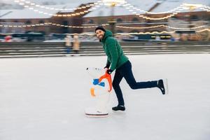 Young male begginer learns skating with help of special figure, leans at it, being on ice rink covered with snow, has cheerful expression, poses in camera. Man uses special equipment for skating photo