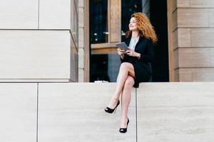 Portrait of elegant gorgeous woman in black suit and high-heeled shoes having slender long legs, sitting with modern tablet, looking aside with happy expression. Female office worker posing outdoor photo