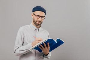 Horizontal shot of concentrated male copywriter writes ideas for new strategy, holds blue notepad, wears spectacles for good vision, holds pencil, stands over grey studio wall with free space for text photo