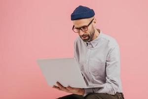 Indoor shot of successful copywriter types publication, reads information, holds laptop computer, wears glasses, shirt, headgear, isolated over pink background with free space for your text or slogan photo