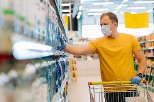 Adult man wears disposable mask and rubber gloves as preventive measures during epidemic time, chooses water in retail store, tries be safe during coronavirus pneumonia outbreak. Self isolation photo