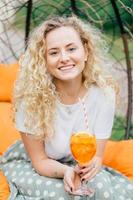 Attractive young female with charming smile, has wavy hair, dressed casually, holds fresh summer cocktail, recreats in garden during weekend. People, rest and lifestyle concept. Vertical shot photo