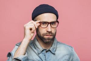 Handsome bearded man with serious expression wears spectacles and glasses, dressed in denim fashionable shirt, isolated over pink background. People, facial expressions and emotions concept. photo