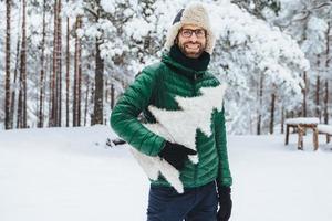 el hombre barbudo de aspecto agradable usa ropa abrigada, sostiene un abeto blanco, se para contra el fondo del bosque invernal, camina con amigos durante el clima helado, está de buen humor. gente y naturaleza foto