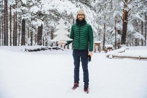 toma al aire libre de un hombre apuesto que usa un sombrero cálido y una chaqueta verde sostiene un abeto de invierno, posa contra árboles cubiertos de nieve, mira felizmente directamente a la cámara. concepto de recreación y temporada foto
