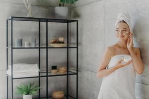 Photo of relaxed young woman applies face cream, has satisfied expression, wrapped in bath towel, poses near grey wall in bathroom, uses cosmetic product for rejuvenation. Beauty and health care