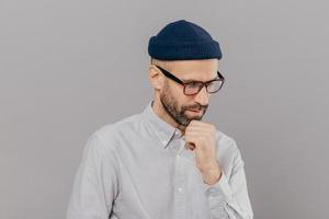 Indoor shot of concentrated man holds chin and focused down, being in deep in thoughts, thinks about innovative idea for his research, has clever expression, wears glasses and white formal shirt photo