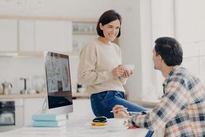 Positive young woman and man discuss renting apartment, pose in coworking space with computer and paper documents, happy wife drinks coffee, looks with pleasant smile at husband, manage budget photo