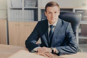 Professional male banker sits in cabinet, wears formal clothes, has confident expression while gives interview concerning banking, poses at office. Man employee arranges meeting of colleagues photo