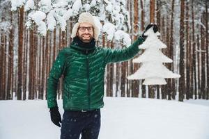 mira este hermoso abeto. alegre hombre barbudo con gorra de piel con orejeras y anorak verde, contento de haber caminado en el bosque de invierno, disfruta de hermosos árboles y paisajes naturales. concepto de temporada foto