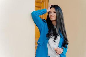 Gorgeous businesswoman with long luxurious dark hair, dressed in formal blue costume and white shirt, has makeup, focused into distance aside, smiles gently, leans with shoulder at white wall. photo