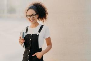 Lovely curly woman with combed hair, dressed in white t shirt, sarafan, keeps hand in pocket, holds book and textbook, wears optical glasses, poses at unversity campus, smiles happily at camera photo