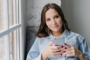 joven mujer de aspecto agradable satisfecha con un buen sonido en los auriculares, sostiene un teléfono celular moderno, se viste informalmente, disfruta del tiempo libre, se sienta en el alféizar de la ventana. mujer escucha canciones de la lista de reproducción en interiores foto