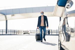 Outdoor shot of adult man returns from business trip, goes to car with suitcase, wears medical mask during coronavirus outbreak, pandemic disease. Passenger arrives after interantional traveling. photo