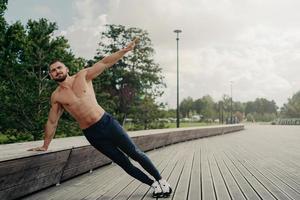 Handsome serious man stands in side plank, has perfect body constitution, poses with naked chest outdoors, keeps arm raised, trains for being healthy, dressed in sport trousers and sneakers.