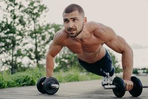 Young handsome bearded sportsman works out outdoor poses in plank with barbells, has strong body, muscular arms. Bodybuilder does exercises in open air, performs weight lifting, push up exercises photo