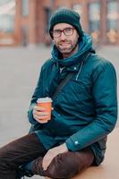 Stylish Caucasian man has free time, poses outside with disposable cup of coffee, dressed in hat and jacket, enjoys good rest, waits for someone at street, has satisfied expression. Lifestyle concept photo