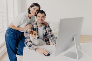 Indoor shot of happy family couple pose near computer monitor, work together remotely, have own business, earn money online, look gladfully at camera, play with favourite pet. People, technology photo