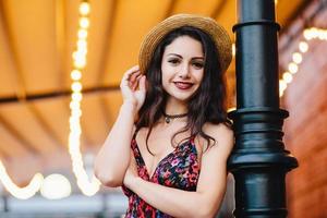 mujer delgada y despreocupada con cabello oscuro y lujoso, ojos brillantes y labios rojos, vestida con sombrero de paja y vestido mientras posa en un café o restaurante. mujer joven feliz descansando en un café al aire libre foto