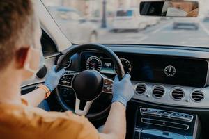 Shot of man driver wears protective medical mask and rubber gloves, poses in car, cares about health during coronavirus. Safety in transport. Health care photo