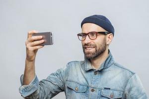 Pleased smiling bearded male with happy expression poses at camera of smart phone, takes photo of himself, wears denim jacket and glasses, isolated over white background. People and technology concept